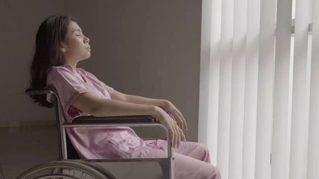 Disabled young woman sitting on wheelchair and looks sad near the window. Shot in 4k resolution