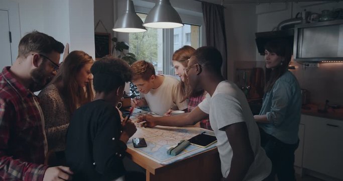 Young casual happy multiethnic student friends using big map on kitchen table with pencils planning travel slow motion.