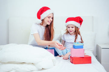 beautiful redhaired ginger woman with santa claus hat and happy little girl in pajamas sitting on bed with white pillow and blanket next to gifts boxes.christmas morning celebration