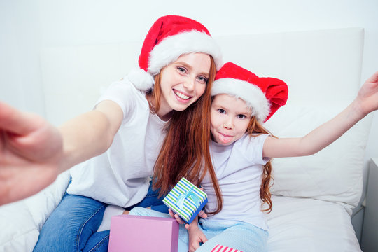 beautiful redhaired ginger woman in santa claus hat and happy little girl in pajamas sitting on bed with gifts boxes,taking selfie pictures on smartphone camera.christmas morning celebration