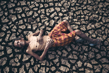 Elderly men lay flat, hands placed on the belly on dry and cracked soil, global warming