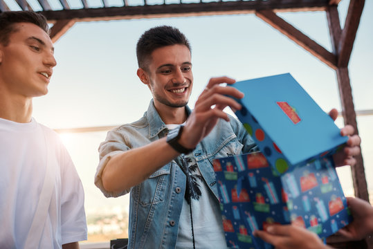 Interesting... Young happy man is opening a gift box while celebrating his birthday with best friends on the roof