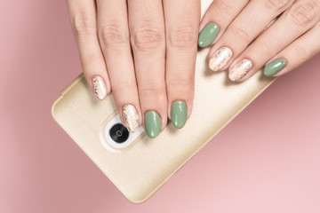 Closeup flatlay top view photography of beautiful two female hands holding modern smartphone in golden case isolated on pink pastel background. Trendy 2 colors green and beige glamourous manicure.