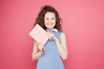 Education concept. Optimistic glad young Caucasian woman with pleased expression,charming smile, blue eyes holds textbooks and pen, rejoices learning new information, isolated on pink wall background