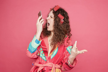Desperate furious housewife gestures with annoyed face expression, talks on smart phone with husband, sorts out relationships, screams loudly, poses against pink studio wall. Negative human emotion.