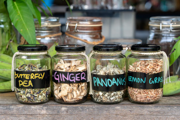 Assortment of dried spices in glass bottles on wooden background. Dry ginger, pandanus, lemon grass and butterfly pea in glass jars, closeup