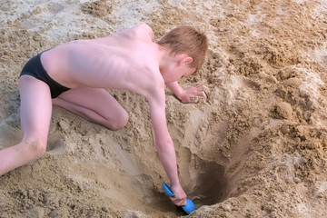 Child boy playing on sand beach digging shovel deep hole and throwing sand on sea ocean coast. Outdoor summer activities at vacation. Boy is sunbathing in swimming trunks.