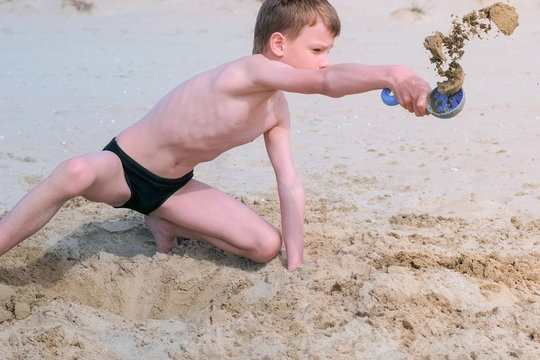 Child boy playing on sand beach digging shovel deep hole and throwing sand on sea ocean coast. Outdoor summer activities at vacation. Boy is sunbathing in swimming trunks.