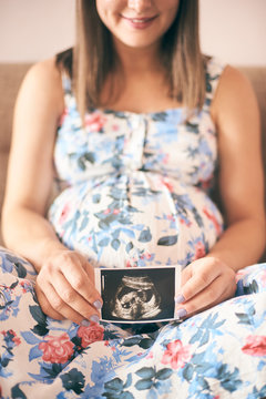 Smiling young woman sitting on sofa and keeping in hands ultrasound image of baby. Cheerful pregnant mother in dress waiting for infant. Concept of new life and expectation.