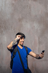 Traveler in blue shirt and backpack with tattoo in the arm, lift sunglasses looking the cellphone with surprise and wow expression in a trip. Oxide and worn wall background.