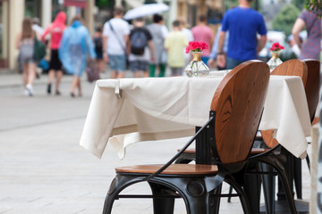 A Table with a white tablecloth small red flowers on it, wooden chair in front, people walking on the sidewalk in the background. Place and setting for a date or a cafe, bistro on the street concept
