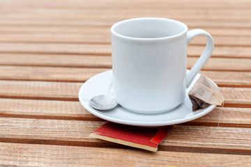 Classy traditional simple single white teacup and a silver spoon laying on a natural wooden table background, wet tea bag on the plate. English tea brewing at the cafe, coffee mug concept, closeup