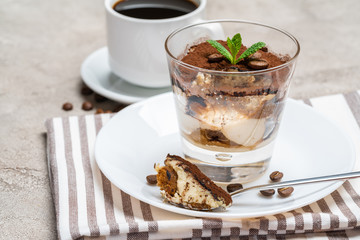 Portion of Classic tiramisu dessert in a glass and cup of coffee on concrete background