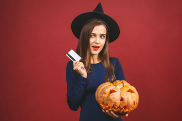 October sale! Halloween Witch with a carved Pumpkin and credit card - isolated on red background. Emotional young woman in Halloween costume. Halloween Party girl.