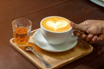 Cup of latte coffee in female hand on white plate on wooden table.