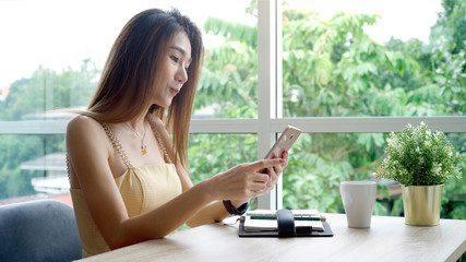 Young beautiful Asian woman enjoying a relaxing moment working and using mobile phone as mobile smart payment to pay for a coffee in the coffeeshop on a bright sunny day