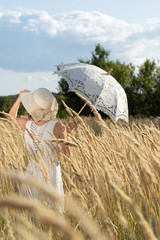 young girl in a field with spikelets holds a lace white umbrella and a straw hat, rear view