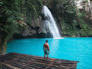 Fit man alone on the bamboo raft in front of the waterfall with turquoise water in Kawasan Falls in...