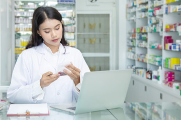 Chemist technician with class dispensing medicine pill on counter with laptop, beautiful Asian pharmacist standing in drugstore doing her work, pharmacist pay attention to the bottle of medicine