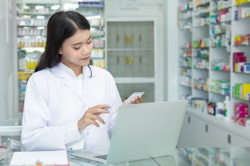 Chemist technician with class dispensing medicine pill on counter with laptop, beautiful Asian pharmacist standing in drugstore doing her work, pharmacist pay attention to the bottle of medicine