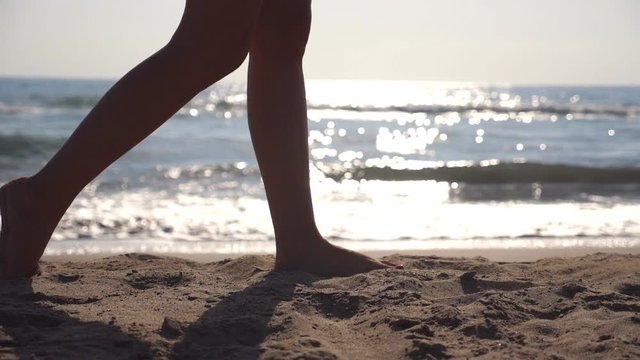 Female feet walking at the sea beach on a sunny day with waves at background. Legs of young woman stepping at the sand. Summer vacation or holiday concept. Side view Slow motion Close up