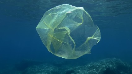  Plastic pollution, Yellow plastic bag in bue water. Slow motion discarded plastic bag slowly drifting under surface of blue water in sunray. Yellow plastic bag floats in Mediterranean Sea, Europe. 