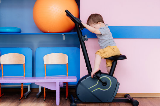 A Little Baby Is Exercising On A Stationary Bike In The Gym