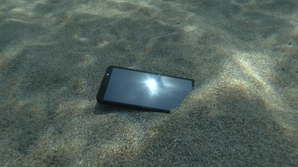 Smartphone lies on a caved seabed on its screen reflects the sun with rays and surface of blue water with small waves. Underwater view. Mediterranean Sea, Europe.