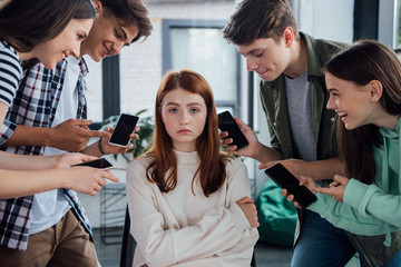 smiling teenagers pointing with fingers at girl during bullying and holding smartphones with blank...