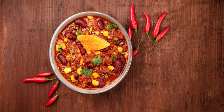 Chili con carne with chilli peppers and a nacho chip, overhead panoramic shot on a dark rustic wooden background with a place for text