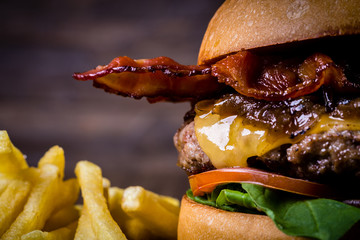 Craft beef burger with cheese, bacon, rocket leafs, caramelize onion and french fries on wood table and rustic background. - obrazy, fototapety, plakaty