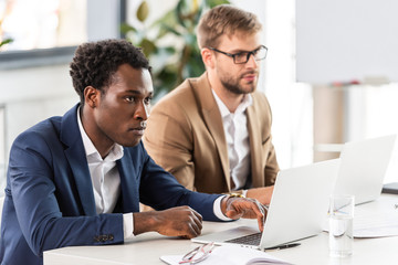 two multiethnic businessmen using laptops at table in office