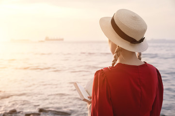An attractive Caucasian girl in a red bright dress and a straw hat sits on a large stone by the sea at sunset and reads an interesting book on the sea horizon. Back view