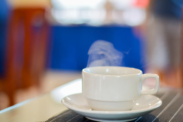 Hot coffee and tea in white ceramic cup on plate with smoke in blurry background.