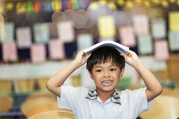 An Asian boy put a book on his head with smiling sitting on a chair in a classroom.