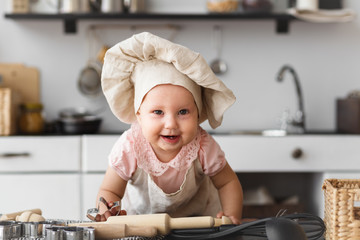 Cute baby cook with wooden tools in a cap and an apron making cookies in the kitchen