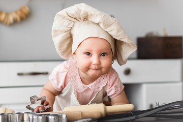 Cute baby cook with wooden tools in a cap and an apron making cookies in the kitchen