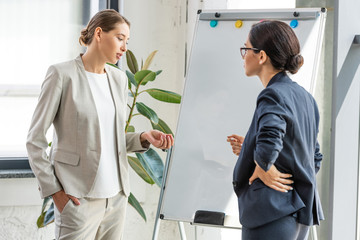 two businesswomen in formal wear standing near flipchart in office