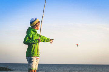 Happy young fisherman with with fishing pole in hand looking the sea on the rocks near the harbour. Sailor boy with waxed jacket enjoying a summer evening. Lifestyle outdoors fun and carefree concept