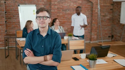 Confident Successful Team Leader Portrait. Happy Young Caucasian Man in Eyeglasses Winks and Smiles with his Diverse Team at Background in Loft Office. 4K Static Medium Shot