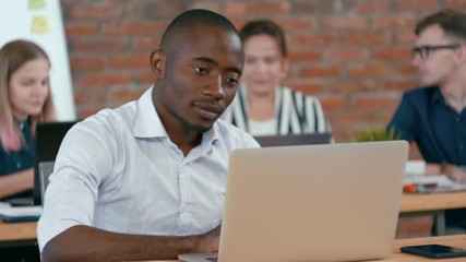 Young African Man is Working on Laptop as Coder or Professional IT Specialist in Authentic Loft Office with Diverse Multiethnic Team. 4K Medium Shot