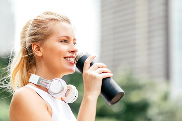 Portrait of motivated woman in sportswear looking away while having break during workout in city park. Happy sportswoman with headphones drinking water after exercising. Horizontal shot. Side view