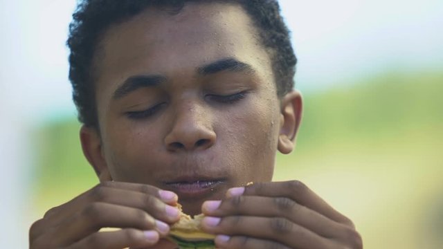Hungry Afro-American teen boy eating delicious sandwich, lunch brake in school
