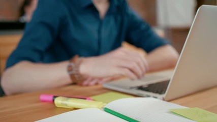 Close up to Male Hand Writing with Pencil in Notebook. Young Man is Working on Laptop in Red Brick Loft Office. 4K Background Universal Shot