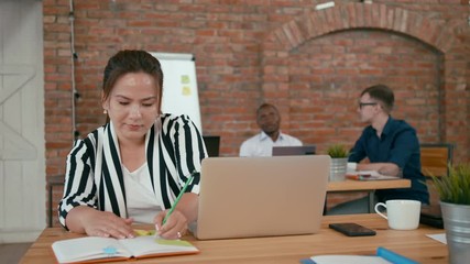 Young Asian Woman is Working on Laptop and Writing by Pencil in Planner in Authentic Loft Office with Diverse Multiethnic Team. Students Discuss Project. 4K Medium Shot