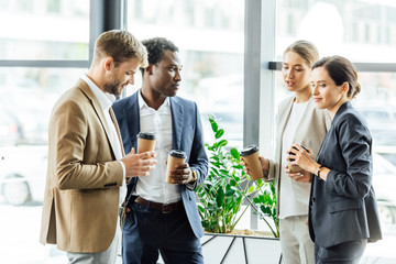 four multiethnic colleagues holding disposable cups of coffee and smiling in office