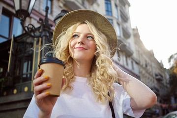 Portrait of pretty smiling blond girl in hat dreamily looking away with coffee to go on city street