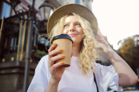 Fuzzy portrait of attractive blond girl in hat dreamily looking away with coffee to go in hand on city street