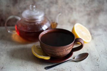 Obraz na płótnie Canvas Cup of tea with lemon on a saucer. Teapot with tea in the background. Hot tea in a clay mug on the gray table.