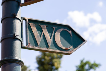 Toilet pointer, wc arrow, restroom icon, green metal sign on pillar in park. Concept sign of toilet room at airport, garden, train station on sky background, outdoor